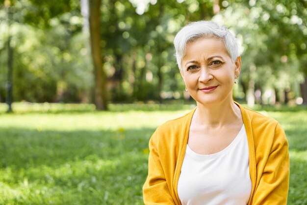 Verano, personas maduras, concepto de edad y ocio. Disparo al aire libre de elegante mujer caucásica pensionista con pelo corto gris vistiendo cardigan amarillo relajándose en la naturaleza salvaje, con una sonrisa