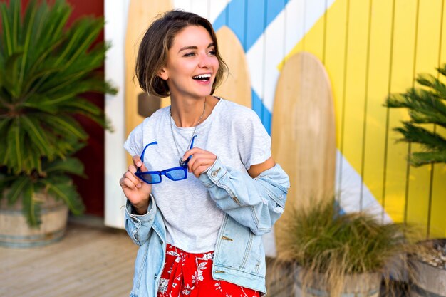 Verano primavera brillante retrato positivo de feliz sonriente mujer morena vestida con traje de moda hipster femenino sonriendo y divirtiéndose, posando frente a tablas de surf y palmeras.