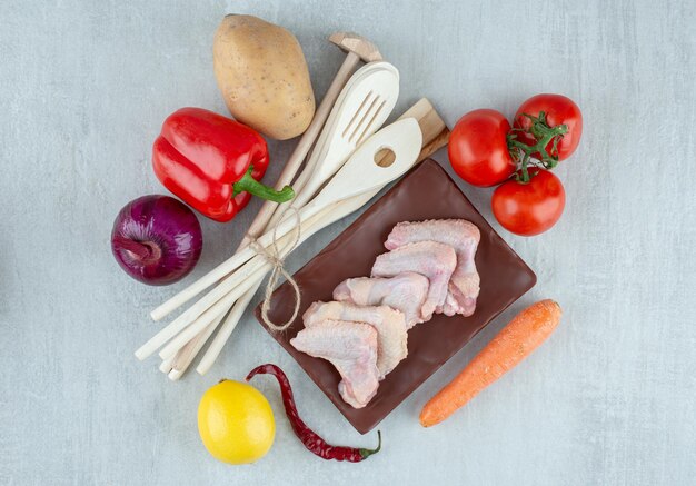 Verduras, utensilios de cocina y alitas de pollo crudo sobre superficie gris.