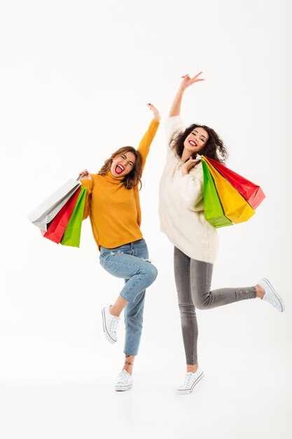 Vertical Dos muchachas alegres en suéteres con paquetes se regocijan sobre la pared blanca