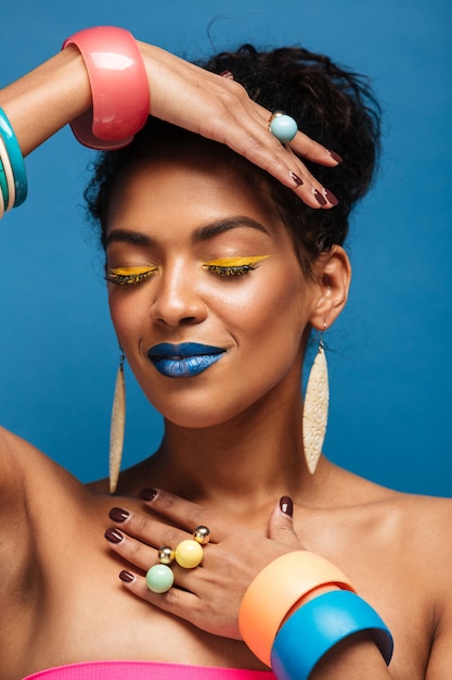 Vertical hermosa mujer afroamericana con cosméticos coloridos cerrando los ojos y gesticulando con las manos aisladas, sobre pared azul