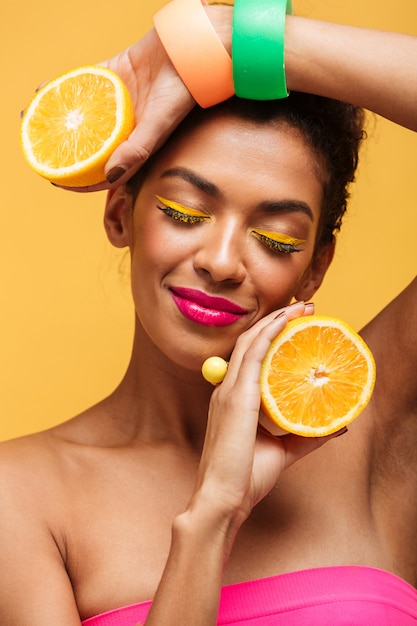 Vertical sensual mujer afroamericana con los ojos cerrados sosteniendo dos partes de naranja y disfrutar de cítricos aislado, sobre pared amarilla