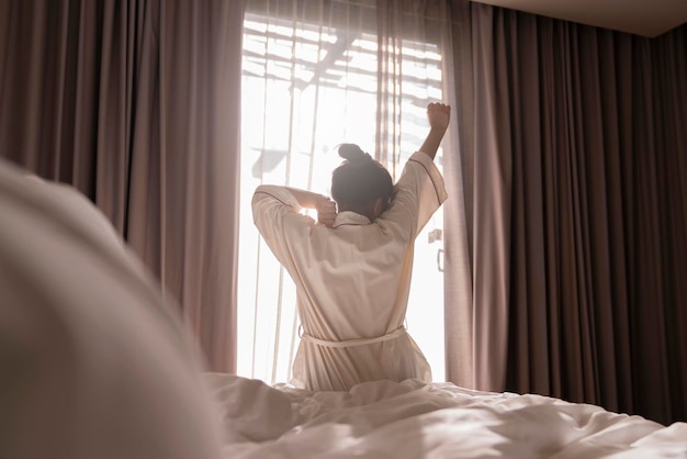 Vestido blanco asiático mujer hermosa mujer estirando mañana despierta frescura y despreocupado momento de fin de semana en un acogedor dormitorio interior con cortina blanca y brillo de sol concepto de hogar de estilo de vida de fondo