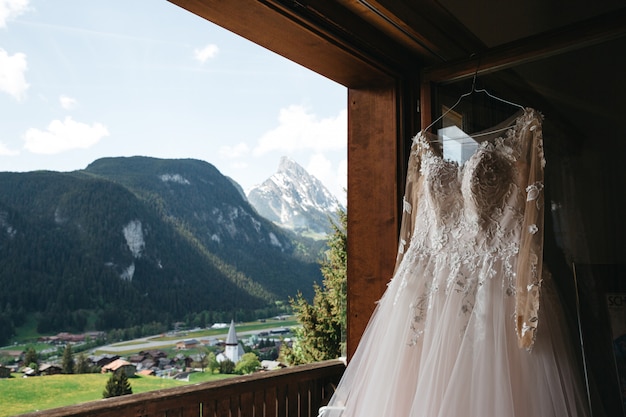 Vestido de novia cuelga de una percha en una ventana con vista a las montañas