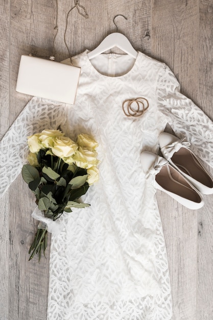 Vestido de novia de novia con embrague de zapatos; cintas para el pelo y ramo de rosas atadas con una cinta blanca sobre fondo de madera