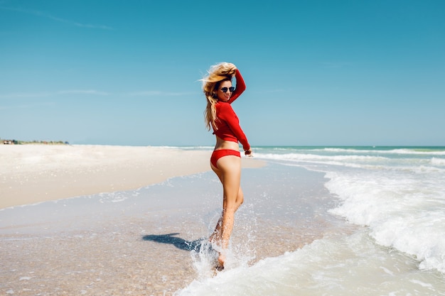 Foto gratuita viajar mujer rubia saltando con expresión feliz, se da la vuelta en el agua. vistiendo bikini rojo. longitud total. vacaciones de verano.