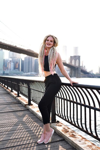 Viaje de mujer joven en Estados Unidos, posando en Nueva York, vista increíble sobre el puente de Brooklyn y la isla de manhattan, viajero urbano con estilo, cabellos rubios inusuales, cuerpo deportivo, viaje a américa.