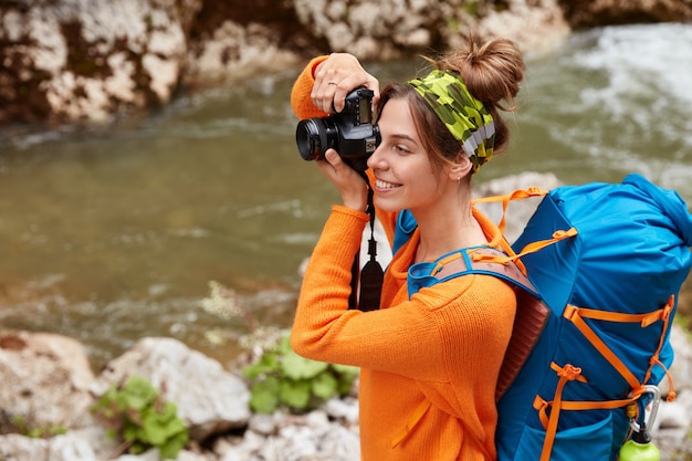 Una viajera alegre toma una foto con la cámara, enfocada en la distancia, usa diadema, suéter naranja, admira la vista panorámica de la naturaleza