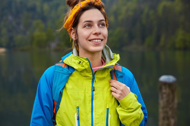 La viajera positiva soñadora tiene recreación activa, posa en el bosque verde y el lago, usa ropa informal