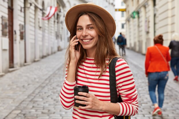 El viajero feliz llama al operador a través del teléfono celular, escucha los beneficios de Internet, se encuentra al aire libre contra el fondo borroso de la calle