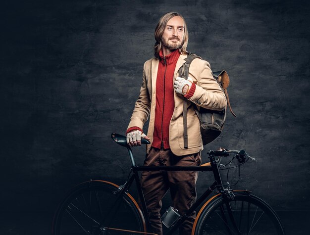 Un viajero hipster con el pelo largo y rubio, con una chaqueta amarilla posando con una auténtica bicicleta de una sola velocidad.