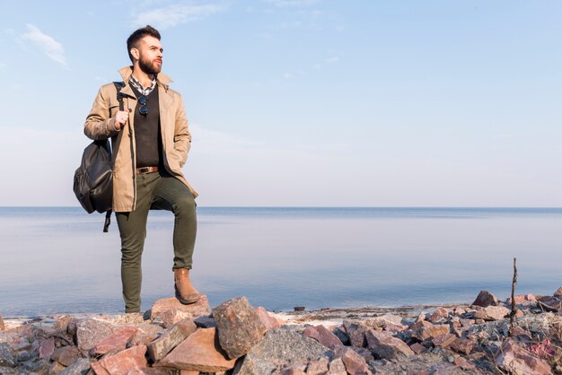 Viajero masculino hermoso que se coloca delante del mar que sostiene el bolso en el hombro que mira lejos