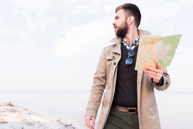 Viajero masculino que se coloca en la playa que sostiene el mapa en la mano que mira lejos