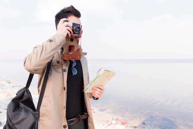 Un viajero masculino que sostiene el mapa en la mano que toma la foto en cámara en la playa