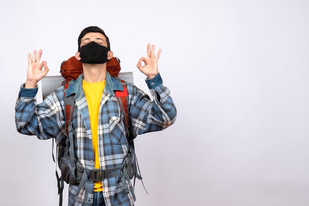 Viajero masculino de vista frontal con mochila y máscara haciendo el signo de ok con los ojos cerrados
