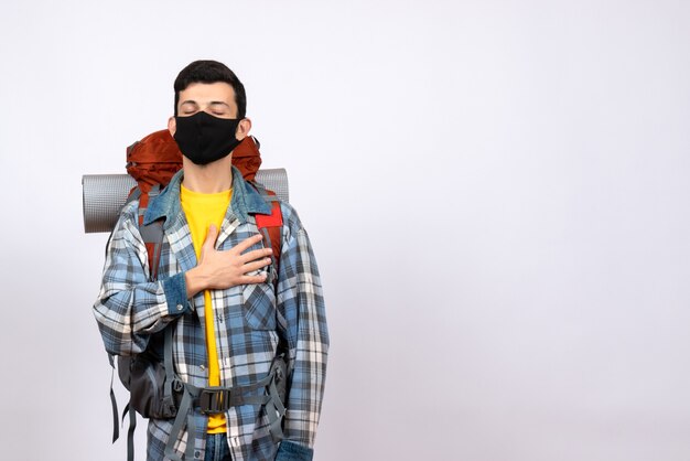 Viajero masculino de vista frontal con mochila y máscara poniendo la mano en el pecho con los ojos cerrados