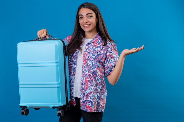 Viajero mujer de pie con maleta de viaje con una sonrisa en la cara que presenta con el brazo de la mano en azul aislado