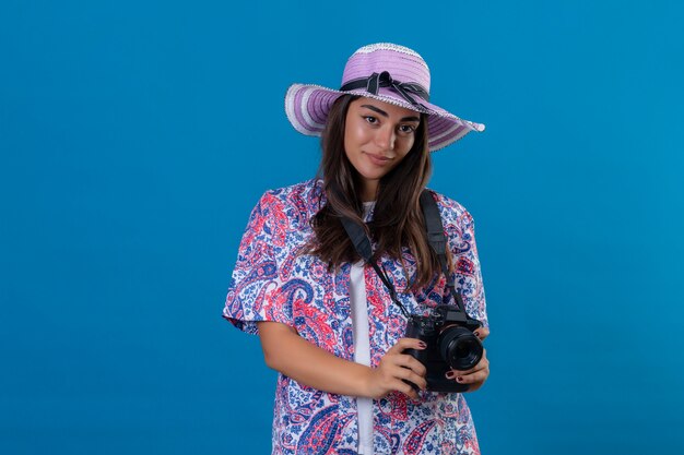 Viajero mujer con sombrero con cámara de fotos con una sonrisa en la cara positiva y feliz de pie en azul aislado