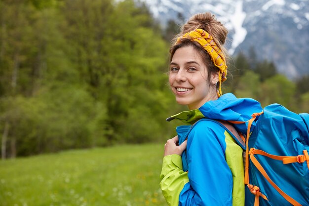 Viajero satisfecho con una sonrisa amable, viste diadema y anorak amarillos, lleva una mochila grande