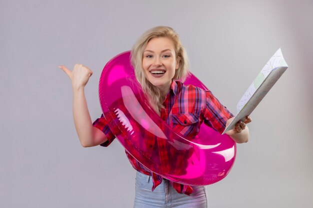 Viajero sonriente joven vestida con camisa roja en anillo inflable sosteniendo el mapa apunta al lado sobre fondo blanco aislado