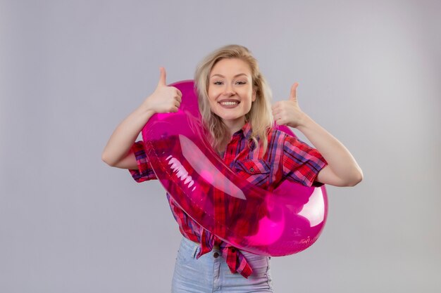 Viajero sonriente joven vestida con camisa roja en anillo inflable sus pulgares hacia arriba sobre fondo blanco aislado