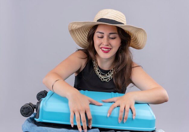 Viajero sonriente joven vestida con camiseta negra con sombrero intenta cerrar la maleta sobre fondo blanco.