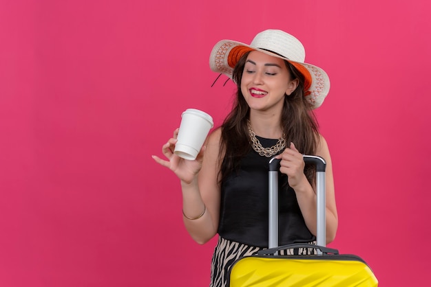 Foto gratuita viajero sonriente joven vestida con camiseta negra con sombrero sosteniendo la maleta y mirando una taza de café sobre fondo rojo.