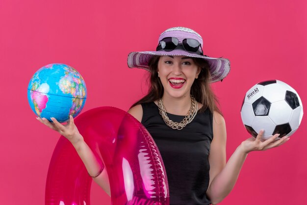 Viajero sonriente joven vistiendo camiseta negra con sombrero sosteniendo círculo inflable y bola con globo sobre fondo rojo.