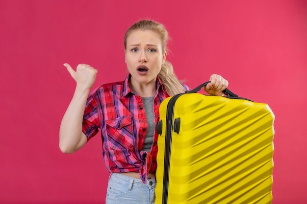 Viajero sorprendido joven vistiendo camisa roja sosteniendo maleta apunta al lado sobre fondo rosa aislado