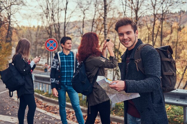 Viajes, autostop, concepto de aventura. Grupo de jóvenes excursionistas parados al margen de la carretera en el hermoso bosque de otoño, tipo con una caminata de planificación de mapas.