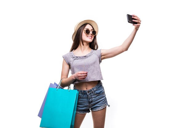 Vibraciones de compras coloridas. Retrato de mujer morena con sombrero y ropa brillante con bolsas de colores tomando selfie con smartphone