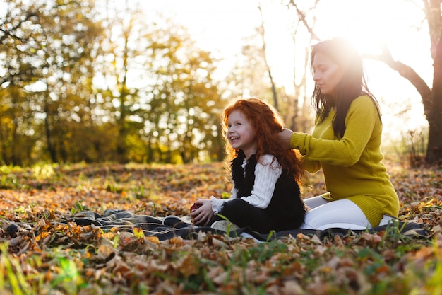 Vibraciones otoñales, retrato de familia. La encantadora mamá y su hija pelirroja se divierten sentadas en el caído