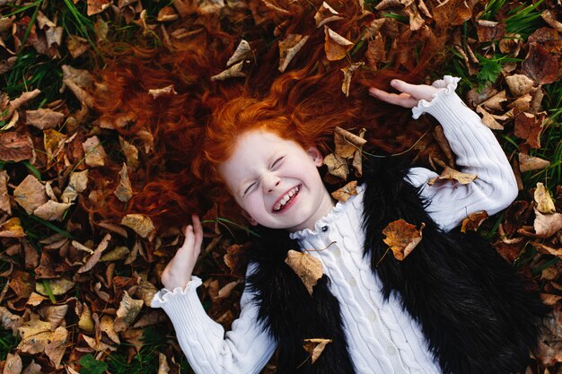 Vibraciones otoñales, retrato infantil. Encantadora y roja niña de pelo se ve feliz acostada en el lecho caído