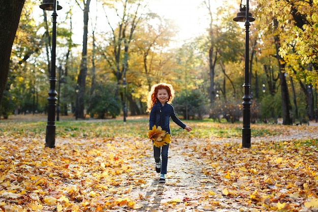 Vibraciones otoñales, retrato infantil. Encantadora y roja niña de pelo se ve feliz caminando y jugando en t