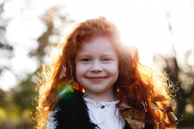 Vibraciones otoñales, retrato infantil. Encantadora y roja niña de pelo se ve feliz de pie en el caído