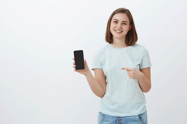 La vida cambió después de este teléfono. Retrato de mujer encantadora encantada de aspecto amistoso con cabello castaño corto en camiseta ligera informal que muestra la pantalla del teléfono celular y apunta al teléfono inteligente sonriendo