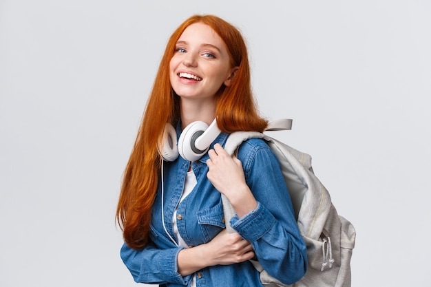 Vida universitaria, estilo de vida moderno y concepto de educación. Estudiante pelirroja guapa alegre con pelo largo astuto, usando audífonos sobre el cuello, mochila, cámara sonriente.