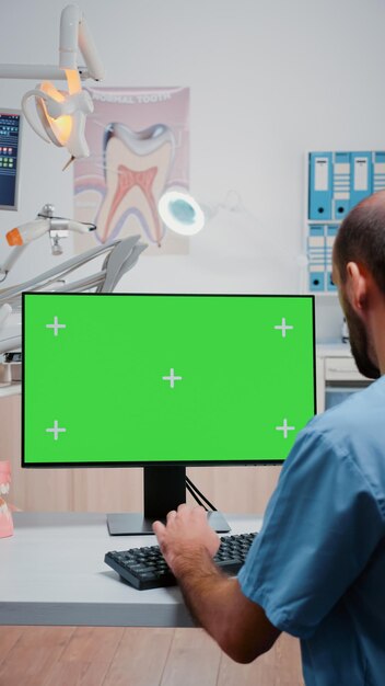 Vídeo vertical: Asistente mirando la pantalla verde horizontal en la computadora en el consultorio del dentista. Hombre que trabaja como especialista en cuidado de los dientes con fondo de maqueta y plantilla aislada en el monitor