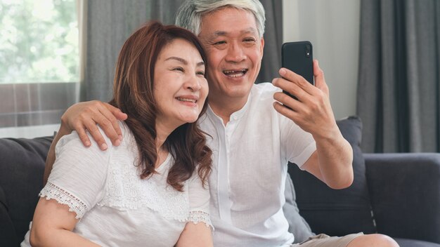 Videollamada asiática mayor de los pares en casa. Abuelos chinos mayores asiáticos, usando la videollamada del teléfono móvil que habla con los niños del nieto de la familia mientras que miente en el sofá en concepto de la sala de estar en casa.