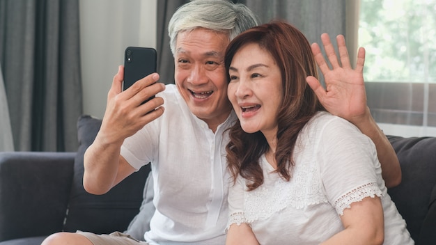 Videollamada asiática mayor de los pares en casa. Abuelos chinos mayores asiáticos, usando la videollamada del teléfono móvil que habla con los niños del nieto de la familia mientras que miente en el sofá en concepto de la sala de estar en casa.