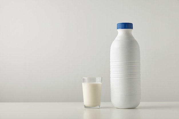 Vidrio transparente con leche orgánica fresca cerca de una botella en blanco de plástico con tapa azul aislada en el lado de la mesa blanca.