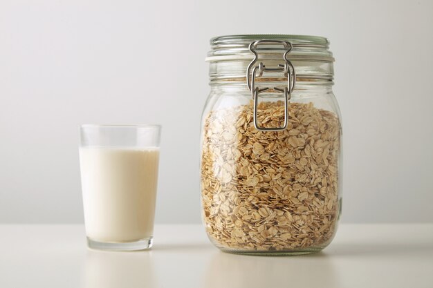 Vidrio transparente con leche orgánica fresca cerca de tarro rústico con copos de avena aislado en el centro de la mesa blanca