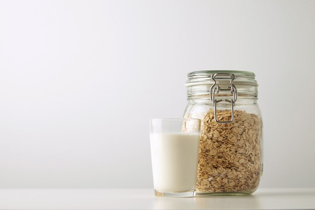 Vidrio transparente con leche orgánica fresca cerca del tarro rústico con copos de avena aislado en el lado de la mesa blanca