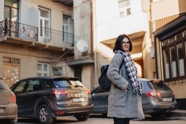 Vidrios que llevan de la chica joven positiva atractiva en una capa en el fondo de edificios en los coches