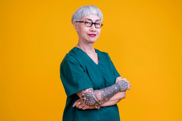 Vieja mujer asiática de cabello gris y tatuaje con anteojos usa uniforme de hospital de pie con una expresión segura y alegre estudio de fondo amarillo