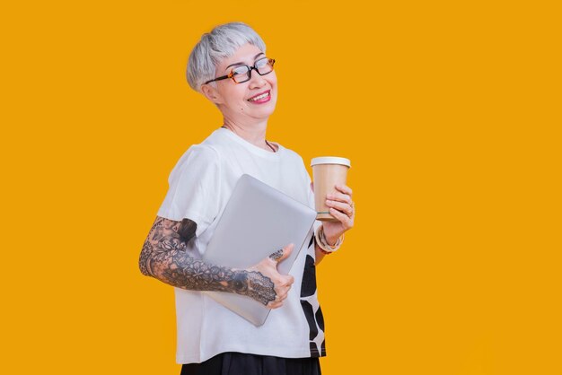 Vieja mujer asiática mayor tatuaje y anteojos propietario de un negocio de tela casual de pie mano asimiento portátil y café sup sonrisa confiada aislar fondo amarillo foto de estudio