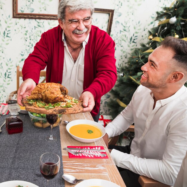 Viejo hombre feliz que pone el pollo asado en la tabla festiva