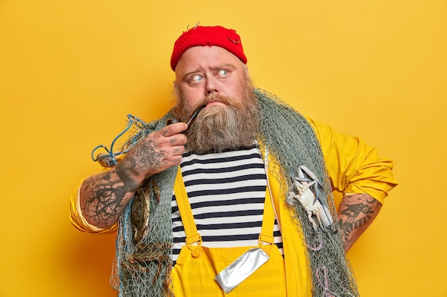 Viejo marinero barbudo experimentado piensa en el día siguiente en el mar, posa con artes de pesca, fuma pipa, vestido con un mono, sombrero rojo