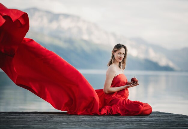 Viento sopla vestido rojo de una mujer embarazada sentada con manzana en el puente sobre el lago