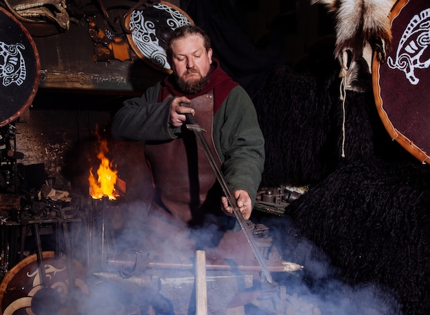 Viking forja armas y espadas en la herrería. Un hombre con ropa de guerrero está en la herrería.